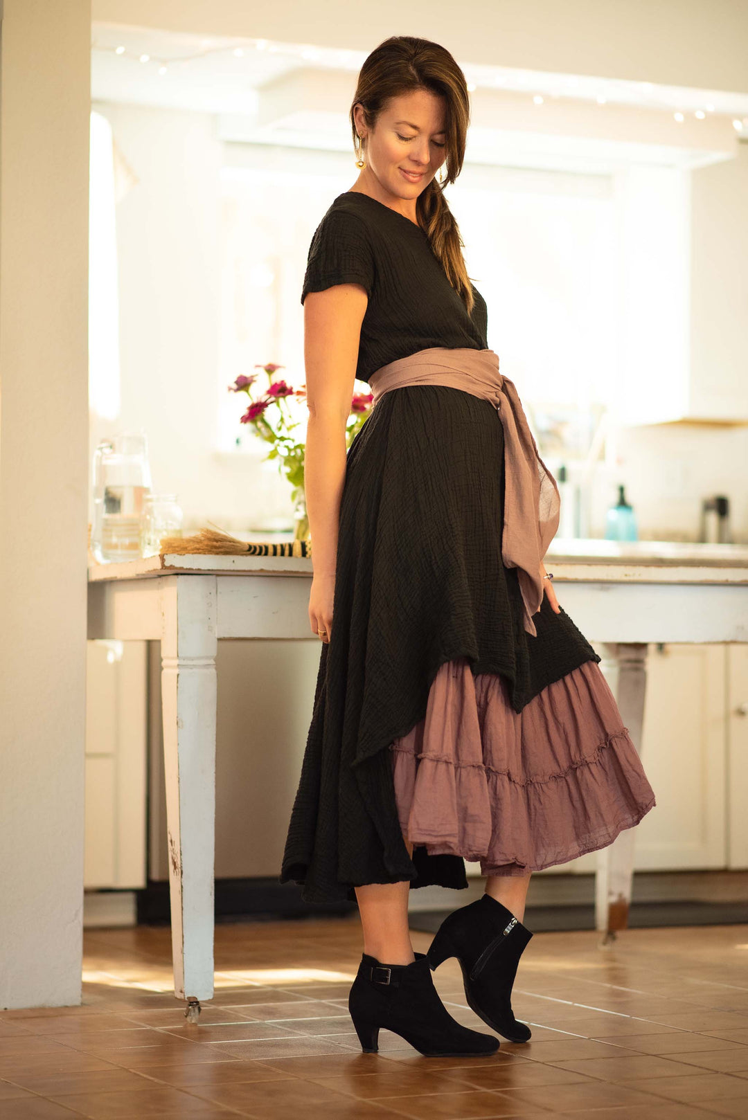 Woman models a long black dress layered over a purple ruffle skirt. She has on a purple belt and black booties.