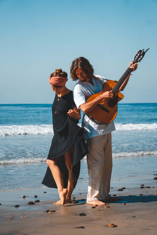 Man and woman stand on beach. Woman is wearing long black gauze cotton dress and pink head scarf. Man is wearing light blue short and khaki pants while holding a guitar.