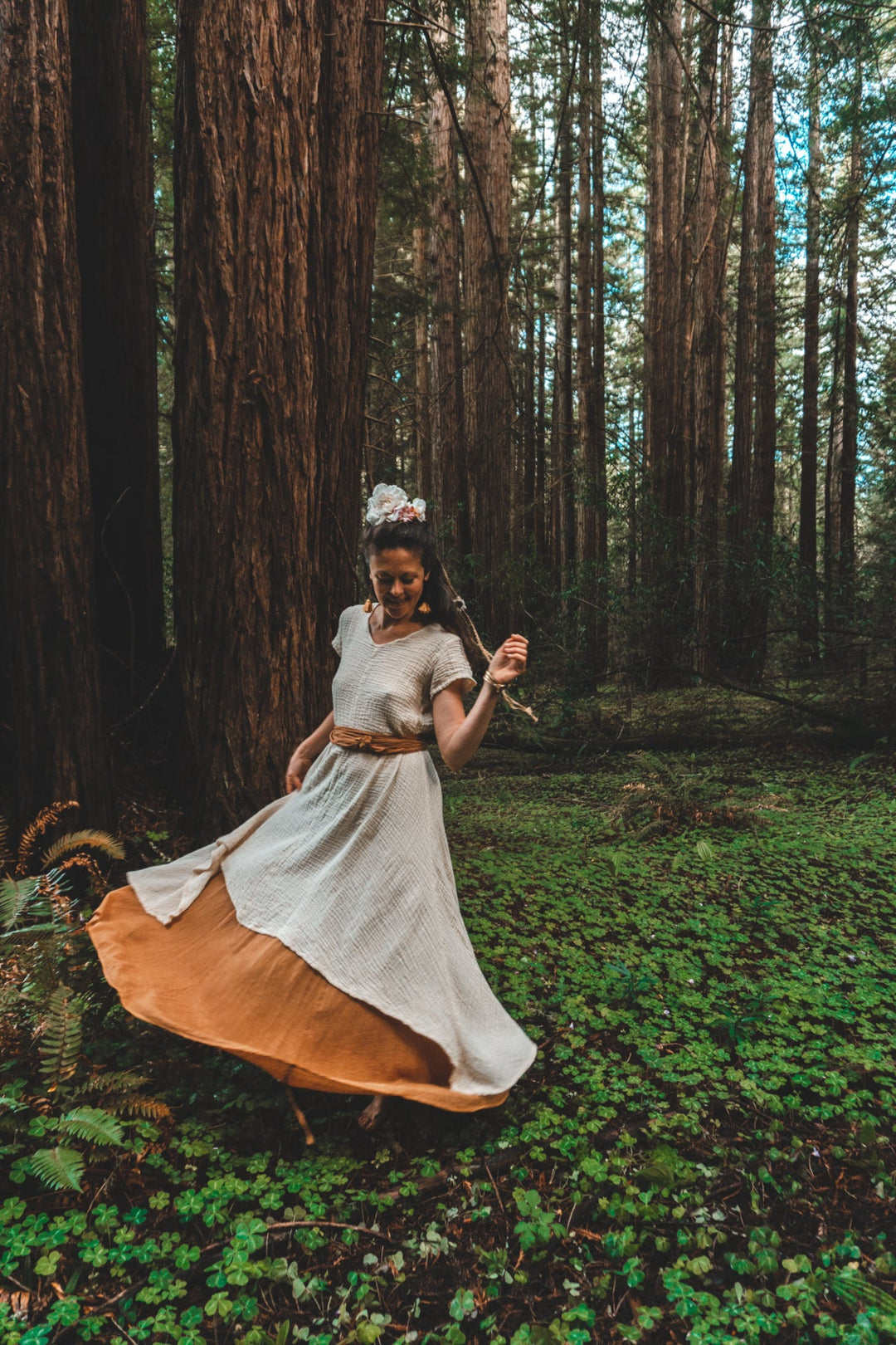 Model twirls in among trees wearing a long dress layered with even longer skirt. She has a belt at her waist.