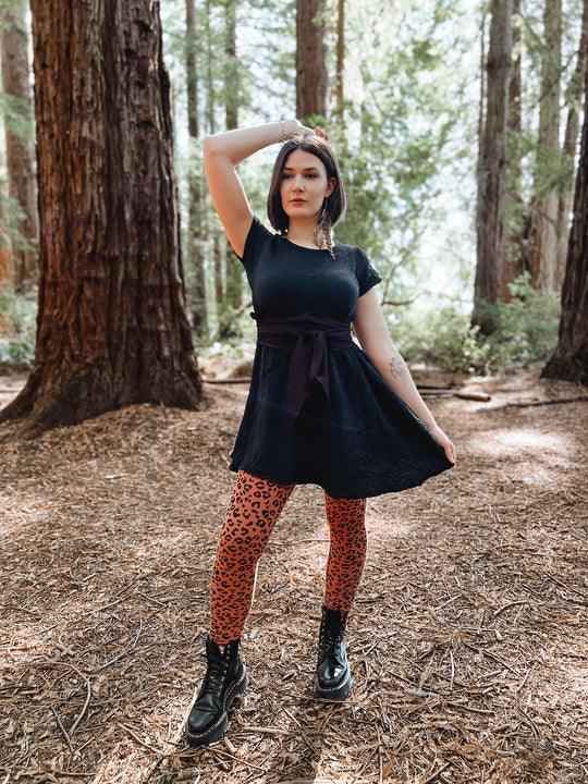 Woman amongst trees is adorned in short dress with sash, leggings and black boots.