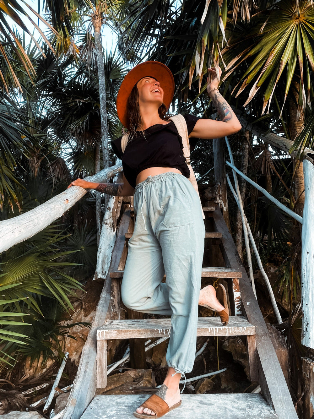 Female model in a hat, black shirt, backpack, blue joggers and sandals walks down outside staircase.