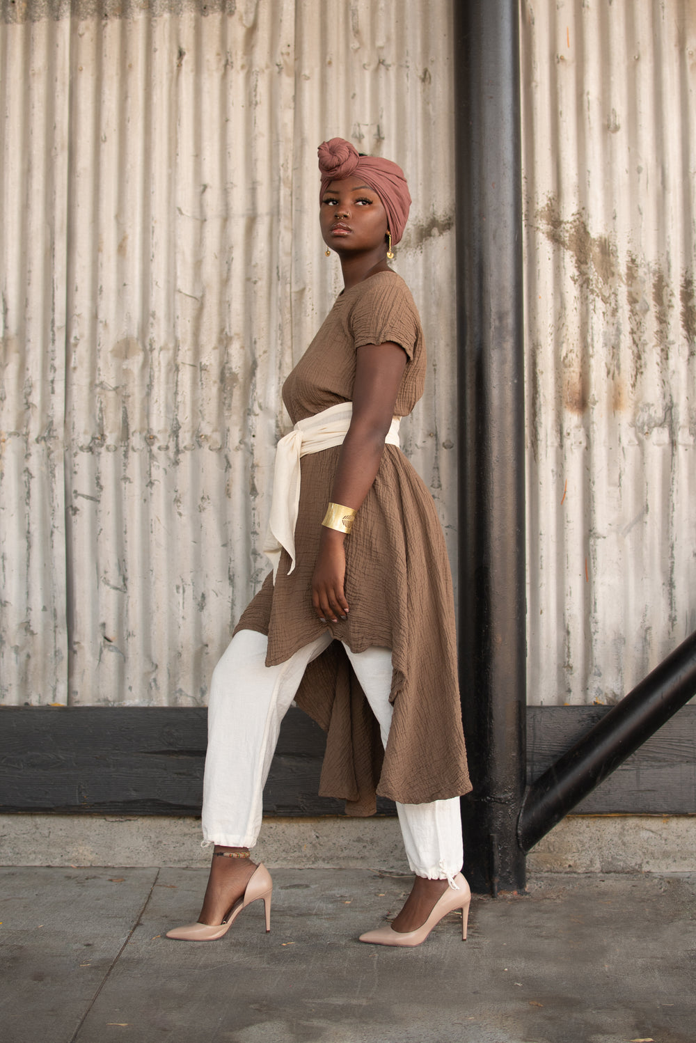 Female model dressed in rose head wrap with long brown tunic top and white bottoms. She has a white wrap around her middle as a sash and tan high heels.