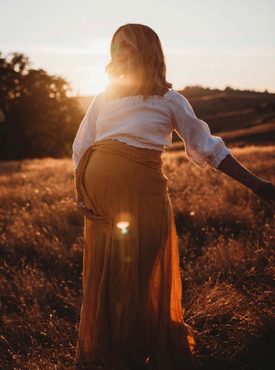 Female model stands outside in a field, lit behind by setting sun. She is pregnant and holding her belly with her right hand. She is wearing a white blouse and gold maxi skirt with bow.