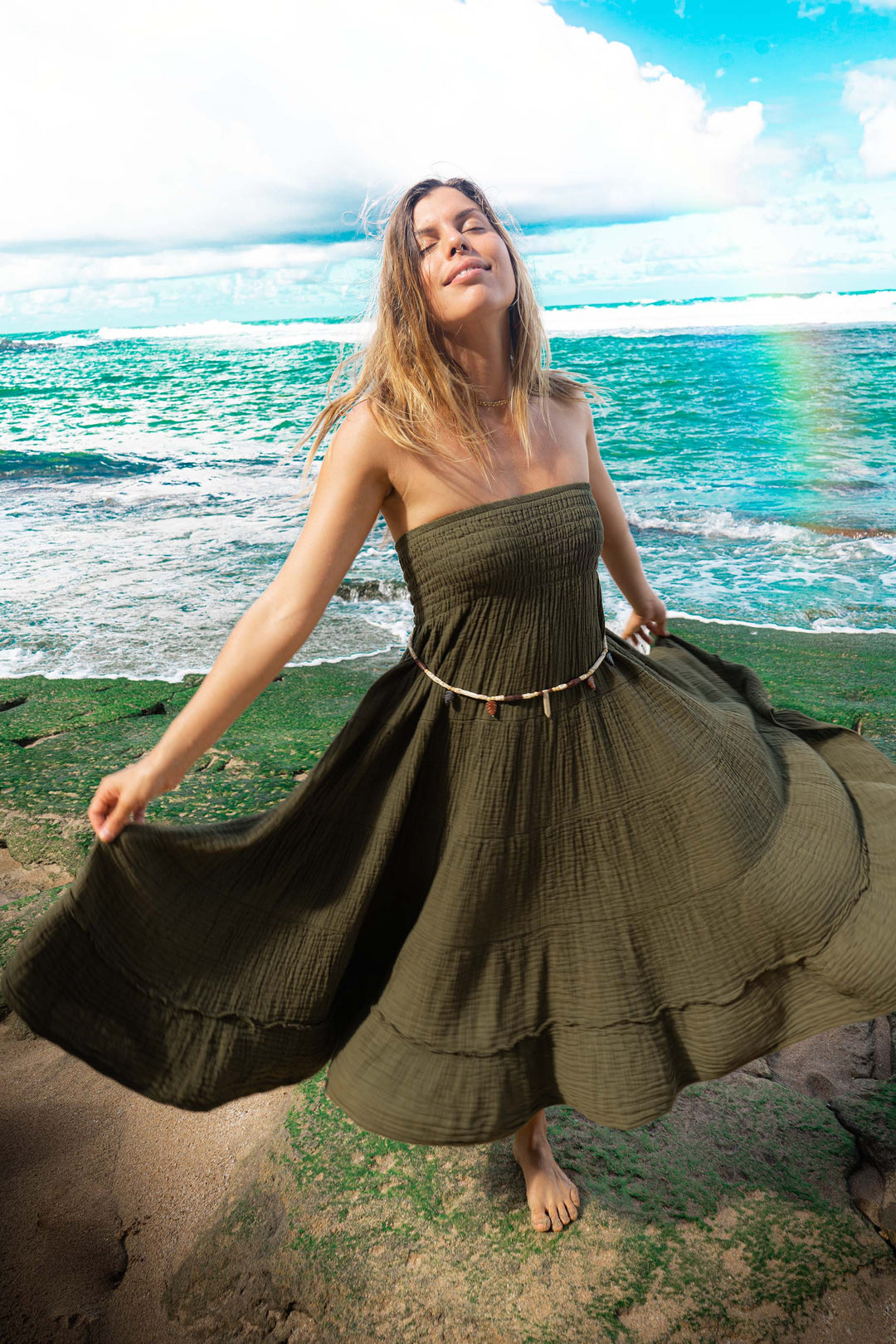 Model wears green strapless dress made of crinkle cotton. She is twirling by the ocean.