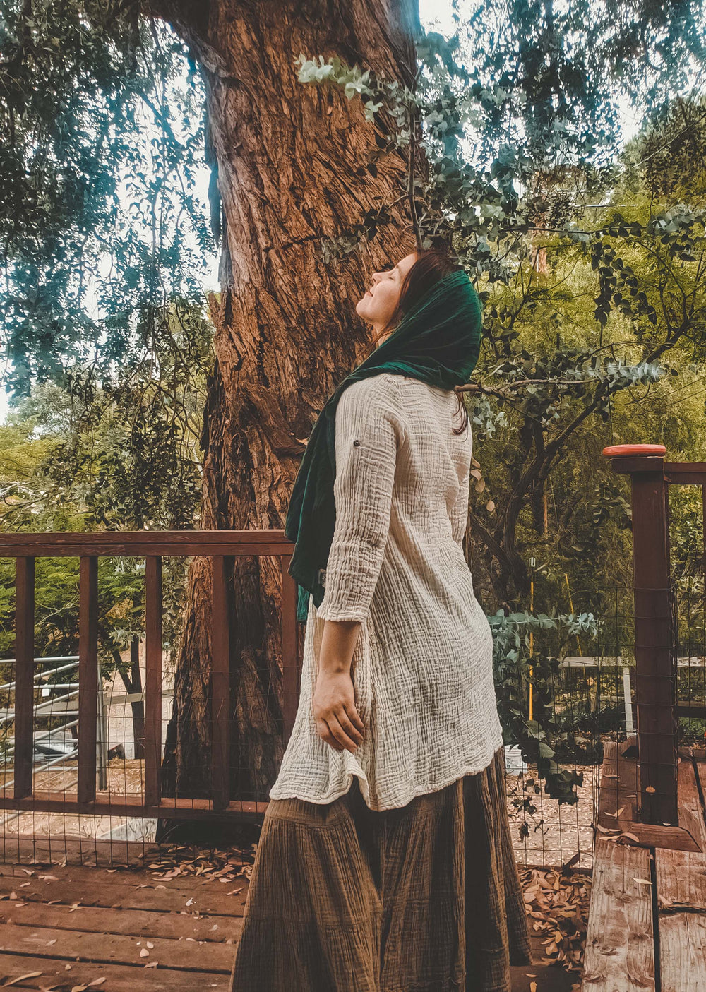 Model has on natural white tunic top over brown maxi skirt. Her hair is covered in a green scarf as she looks to the sky.