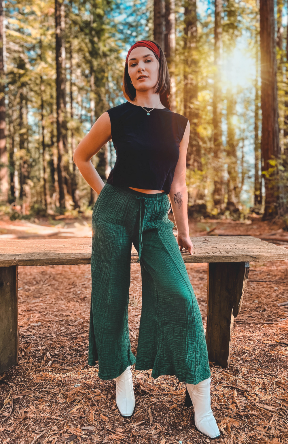 Woman wears black crop tank top with emerald green pants and white boots.