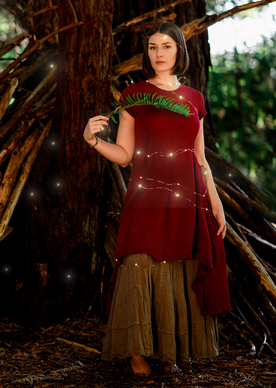 Model is clothed in short-sleeved red dress layered over a long green skirt. She holds the branch of a pine tree.