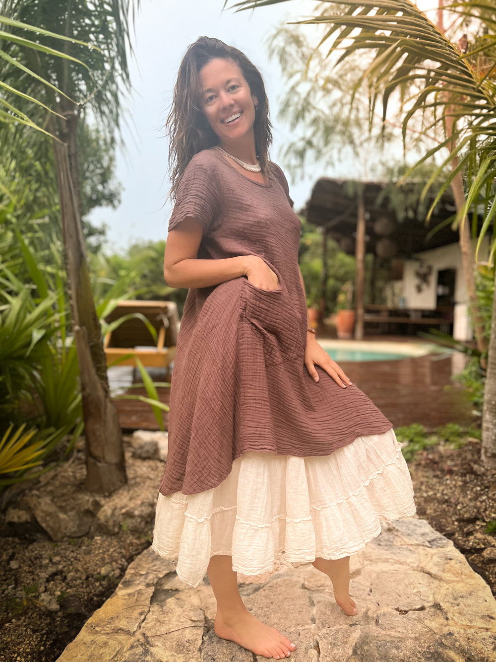 Woman has layered short dress with longer skirt. She is barefoot and smiling.