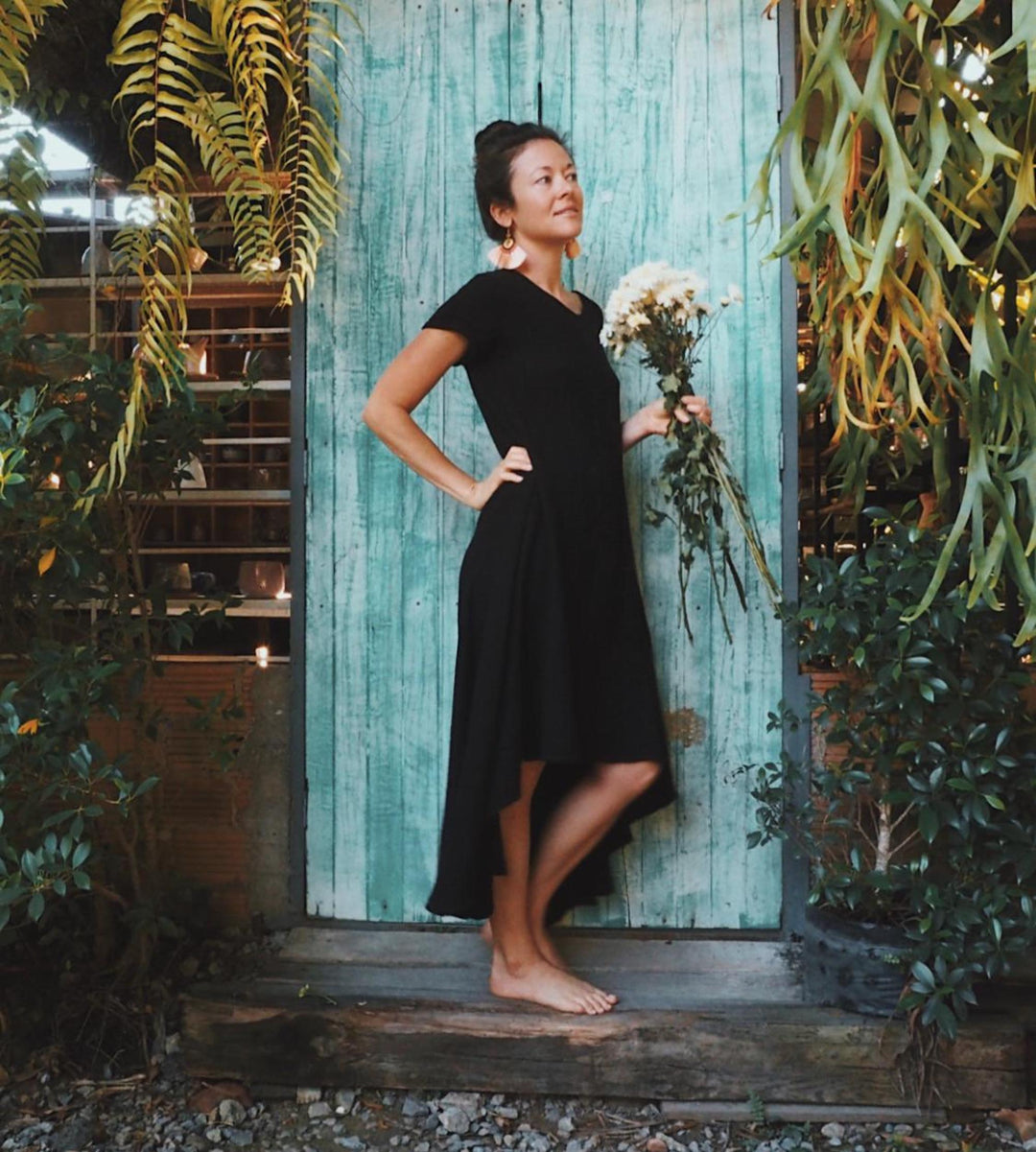 Woman stands in front of a blue door wearing a simple black dress while carrying white flowers.