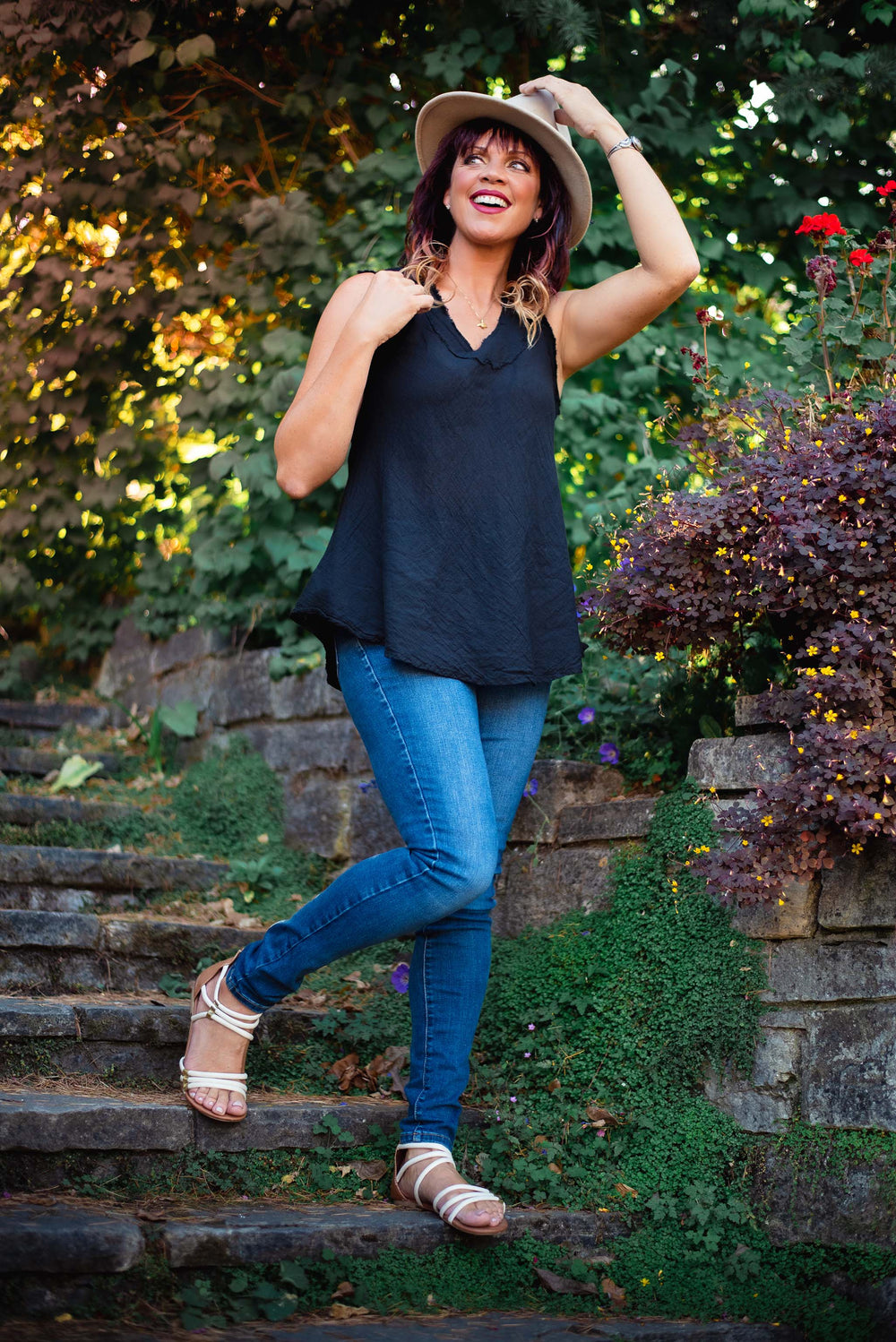 Female model dressed in hat, long black tank top, jeans and white sandals walks down stone stairs in garden.