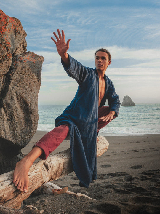 Male model balances in driftwood by the ocean wearing a loose blue robe and red pants.