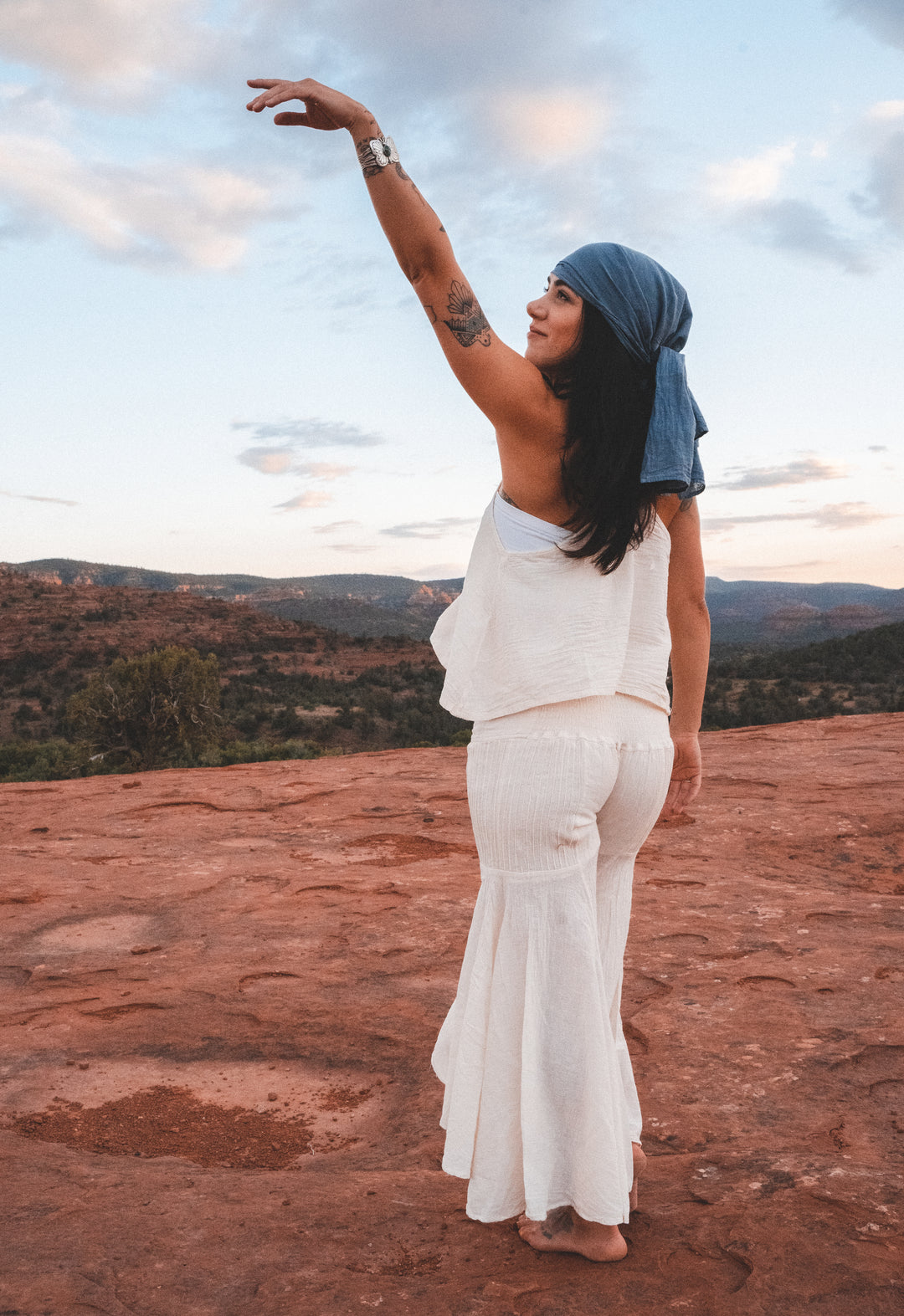 Woman stands outside on red rock wearing a slate blue head wrap, natural tank top and un-dyed lightweight pants. Her left arm is up in the air.