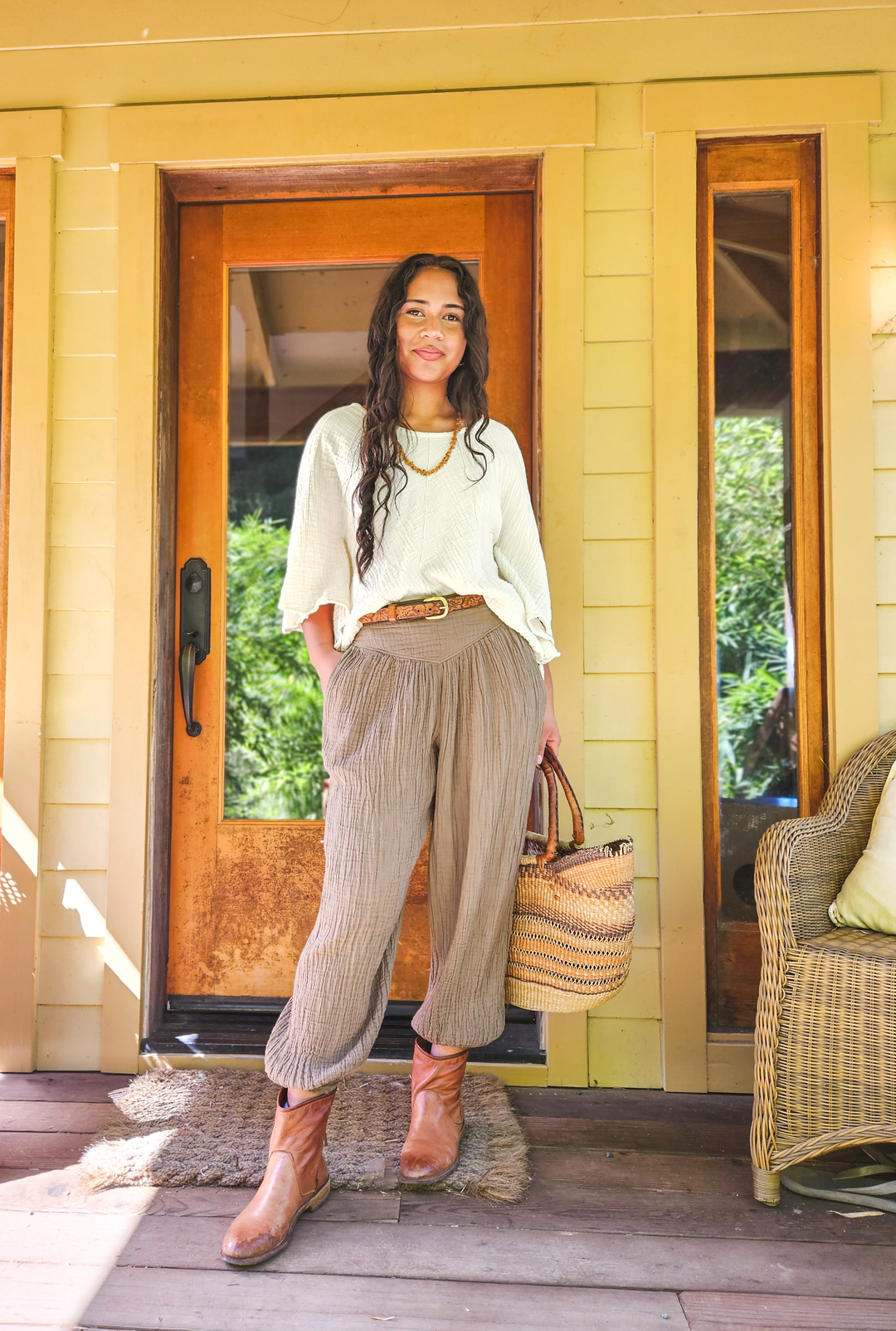 Woman stands on front porch dressed in white gauze shirt paired with long neutral pants belted at waist.