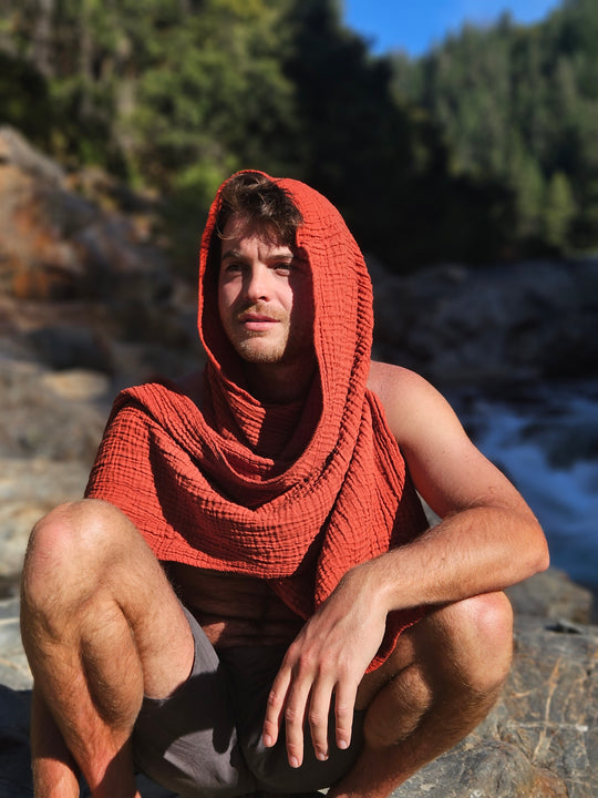 Male model has red gauze scarf draped over head and shoulder. He crouches down on rock.