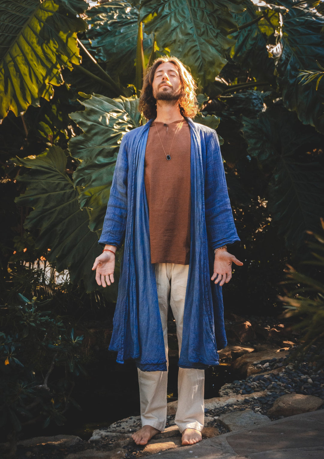Male wears blue robe over brown shirt and white pants. He stands barefoot in the jungle.