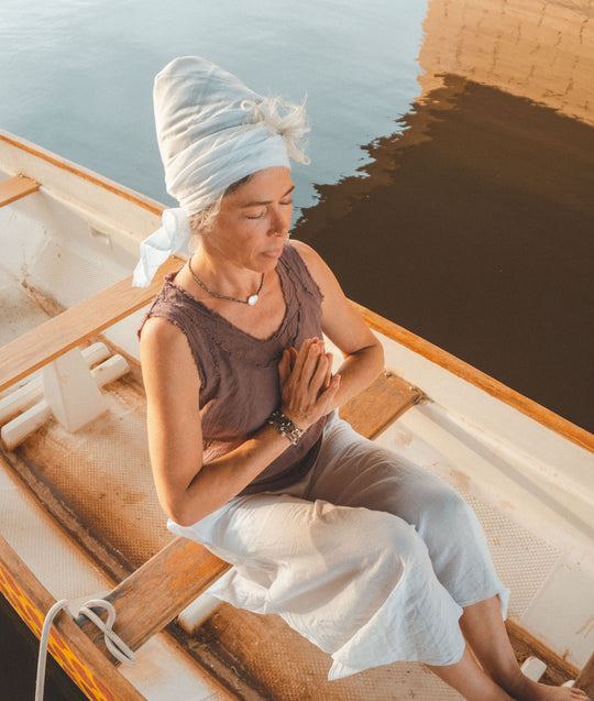 Woman sits on boat wearing white head wrap, brown tank top and loose white pants. Her hands are in prayer pose.