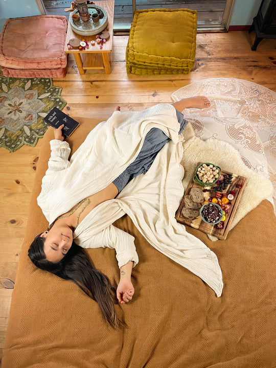 Female lays on bed with tray of food beside her. She is clothed in a natural cloak with blue bottoms.
