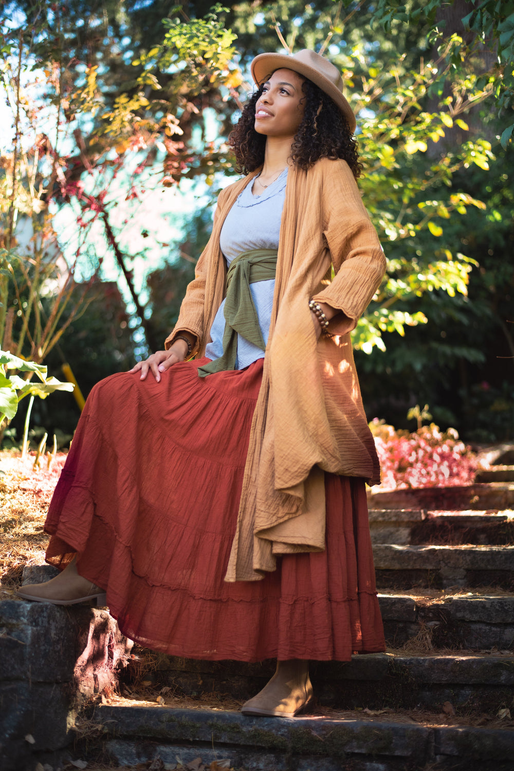 Model poses on stone garden stairs adorned in long yellow cloak, ruffled shirt with belt and maxi skirt.