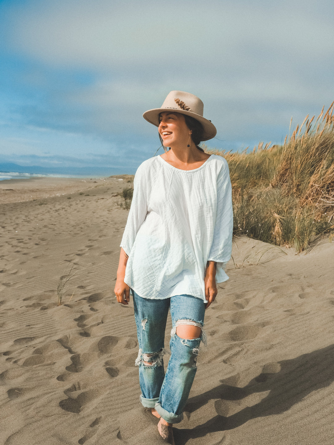 Woman walks barefoot on sand near ocean wearing hat, un-dyed shirt with three quarter length sleeves and jeans.