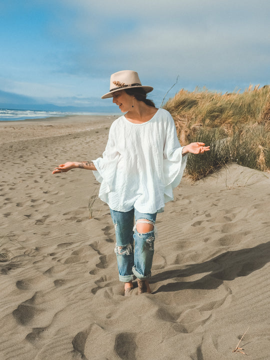 Woman walks barefoot on sand near ocean wearing hat, blousy gauze shirt and blue jeans.
