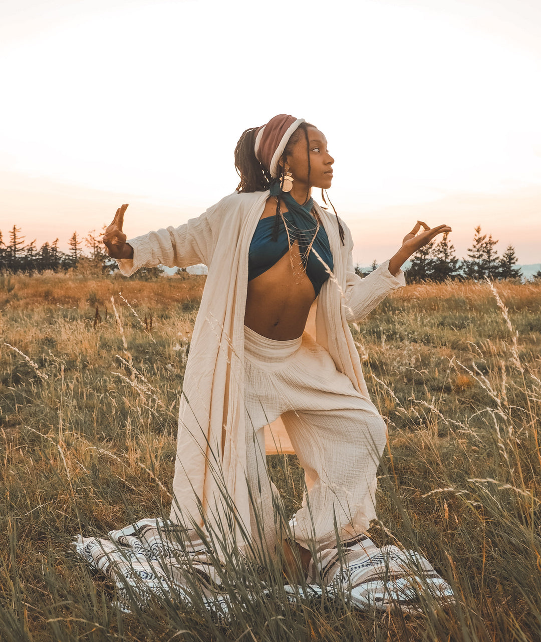 Female model in a yoga pose wears head wrap, scarf top, long white cloak and loose white pants.