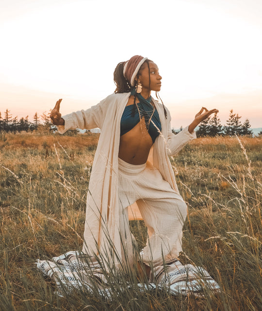 Female model in a yoga pose wears head wrap, scarf top, long white cloak and loose white pants.