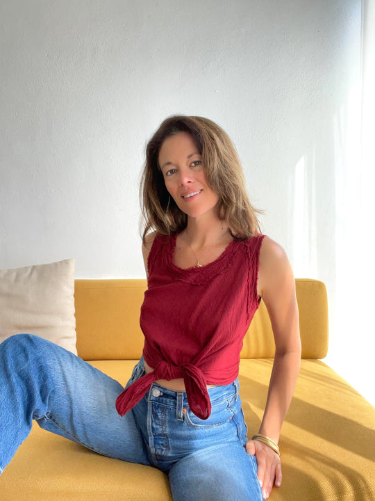 Model smiles on mustard couch wearing red tank top tied at waist and jeans.