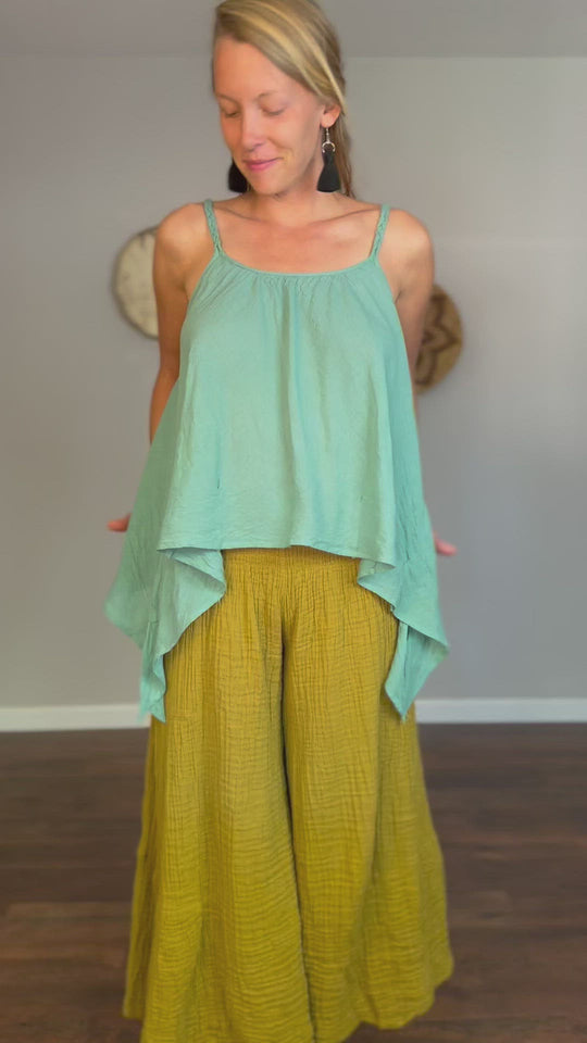 Female clothing model is wearing a aqua blue tank top and olive green gauze pants. She shows how to tie the tank top in a knot at her waist.