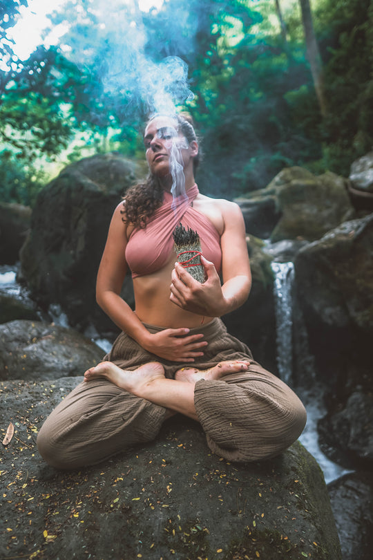 Female model sits in meditation pose on a rock wearing scarf top and loose brown leggings. She is burning incense. 