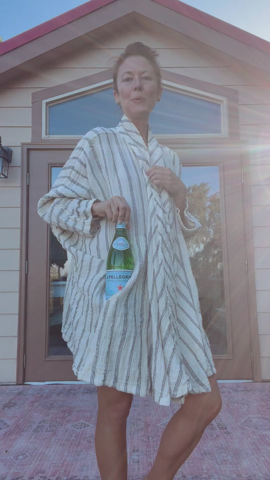 Model is wearing long striped robe with two pockets. She slides a glass bottle into one of the large pockets.
