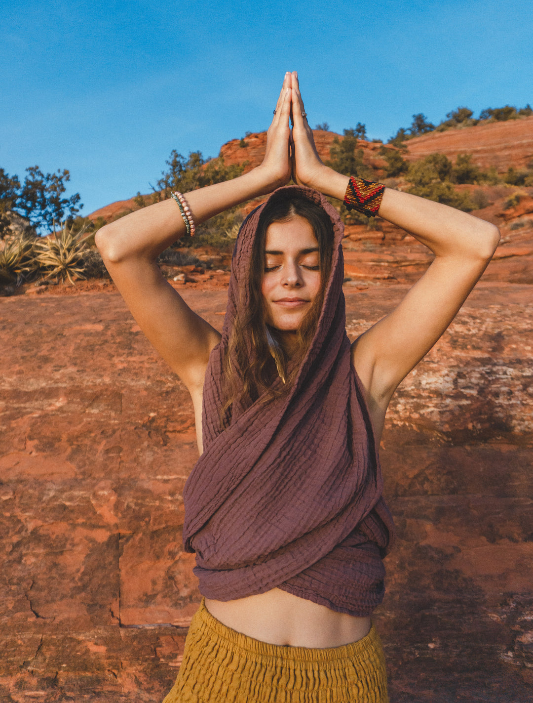 Woman has earthen rose colored scarf wrapped around head and upper body as a top. She has on gold-color bottoms and stands in a yoga pose with hands in prayer above her head.