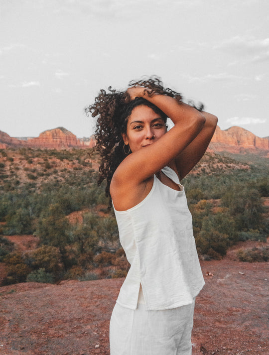 Female model stands with arms up and hands in her curly brown hair. She is wearing an un-dyed tank top and pants. She is standing on red rock with a backdrop of mountains.