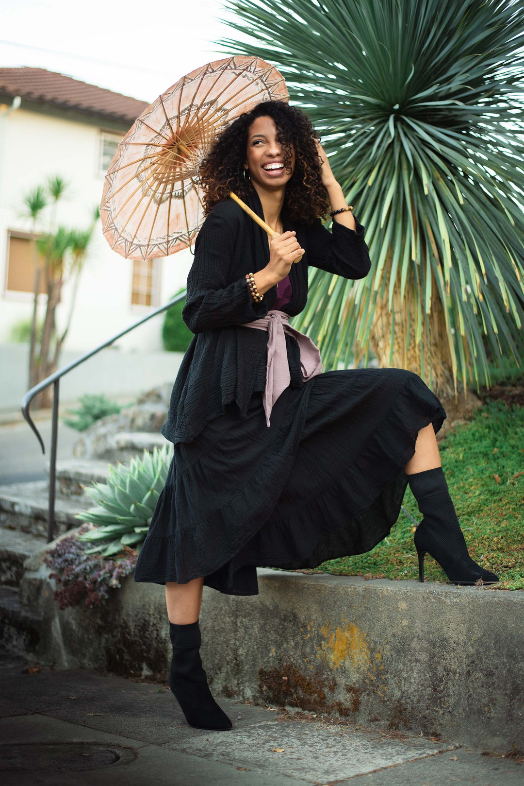 Model dons black wrap and black skirt tied with pink fabric sash. She is carrying a decorative umbrella. 