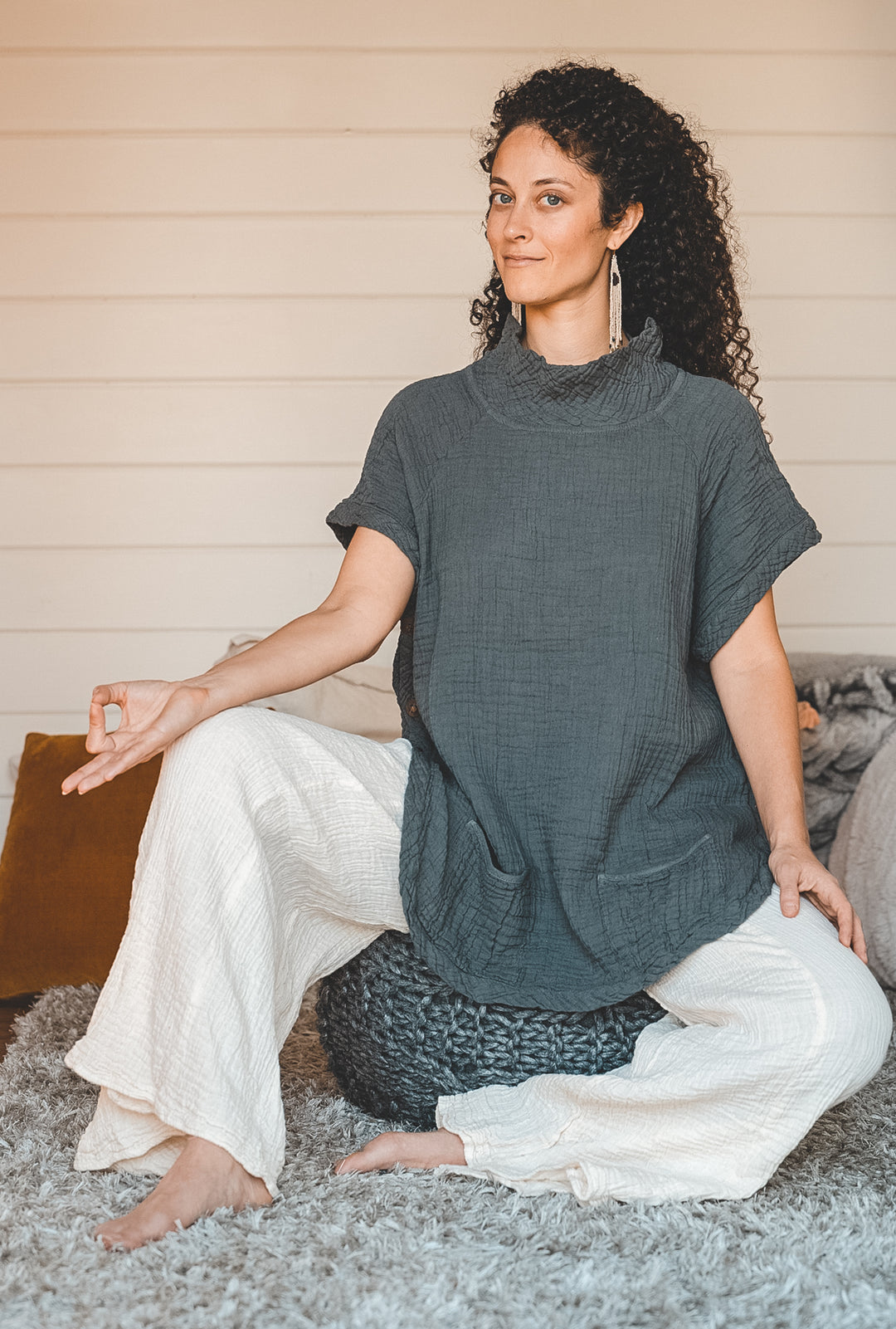 Woman wears gray shirt and white pants while sitting on meditation cushion. 