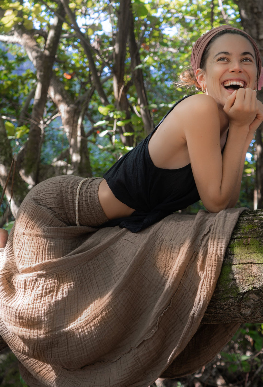 Woman laughs while resting across tree limb wearing long brown gauze skirt.