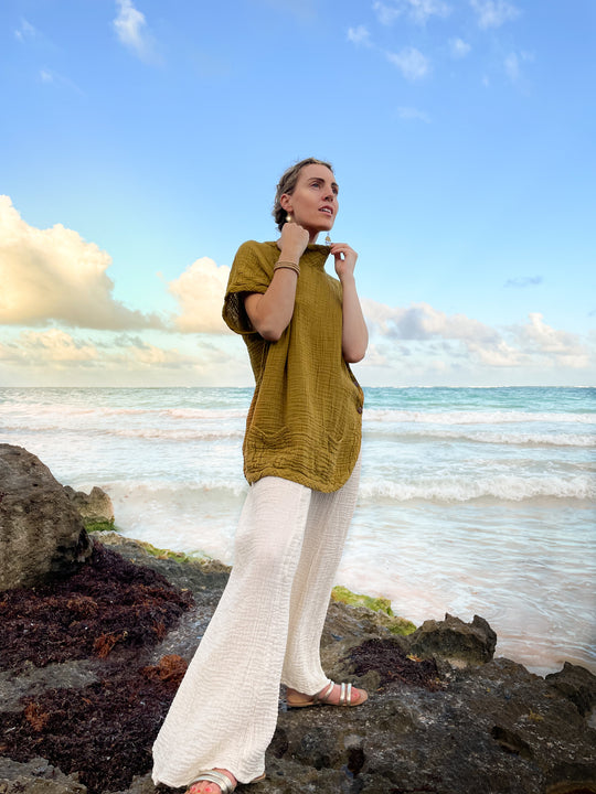 Staring out at the water is a woman in a long gold tunic and white gauze pants.