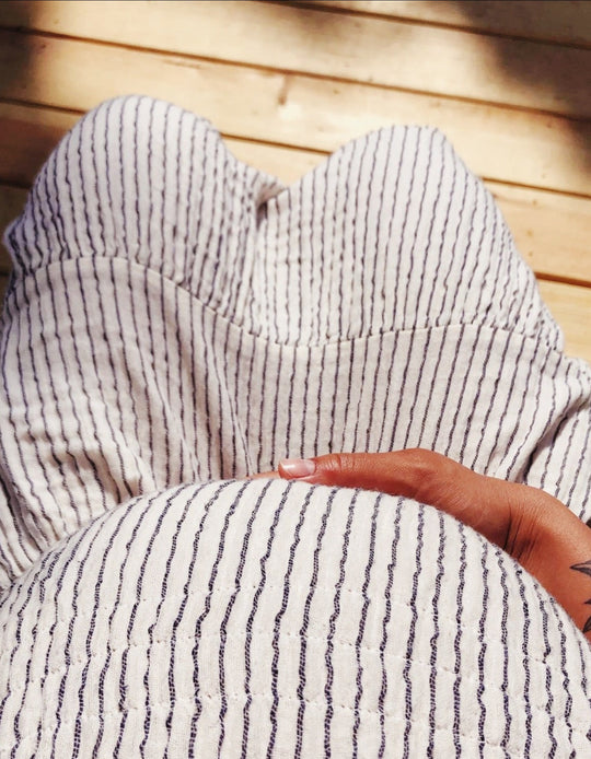 Clothing model is pregnant and wears long white dress with pinstripes while seated. Woman has hand on pregnant belly.