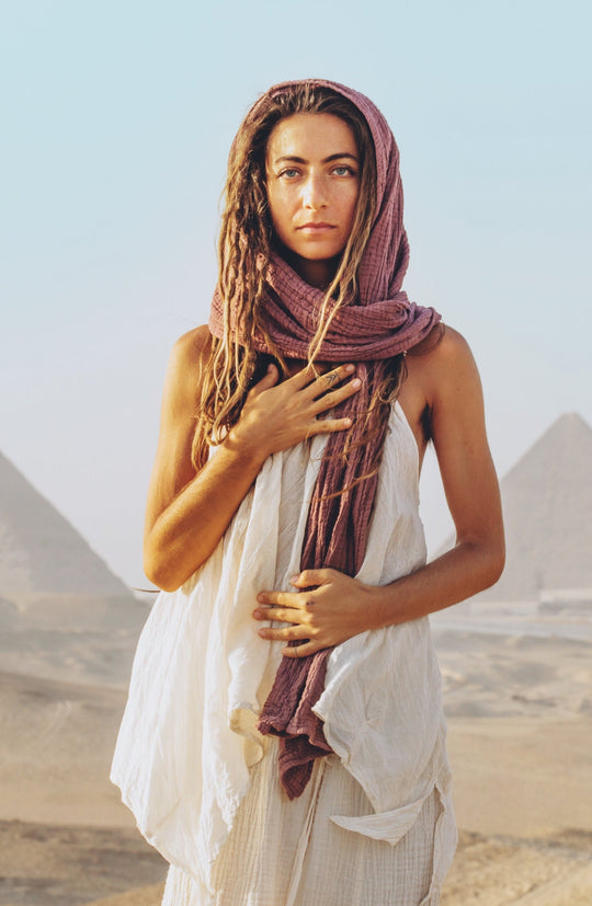 Female model wears rose-color wrap as a scarf on her head paired with white tank top and white bottoms. Her right hand is at her heart.
