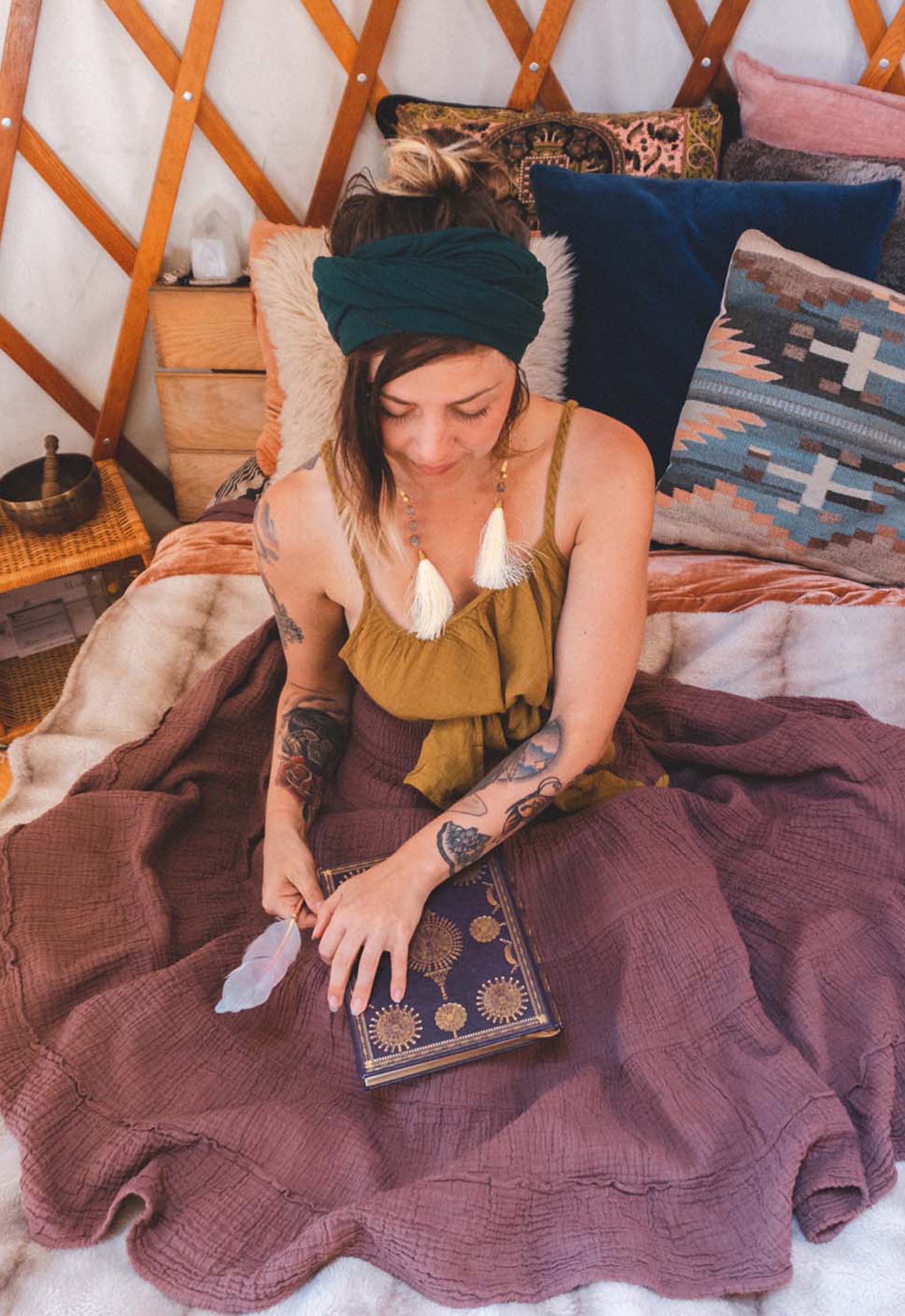 Woman sitting on bed with colorful pillows wearing full, layered purple skirt, gold tank top and green head wrap. She had a book on her lap.
