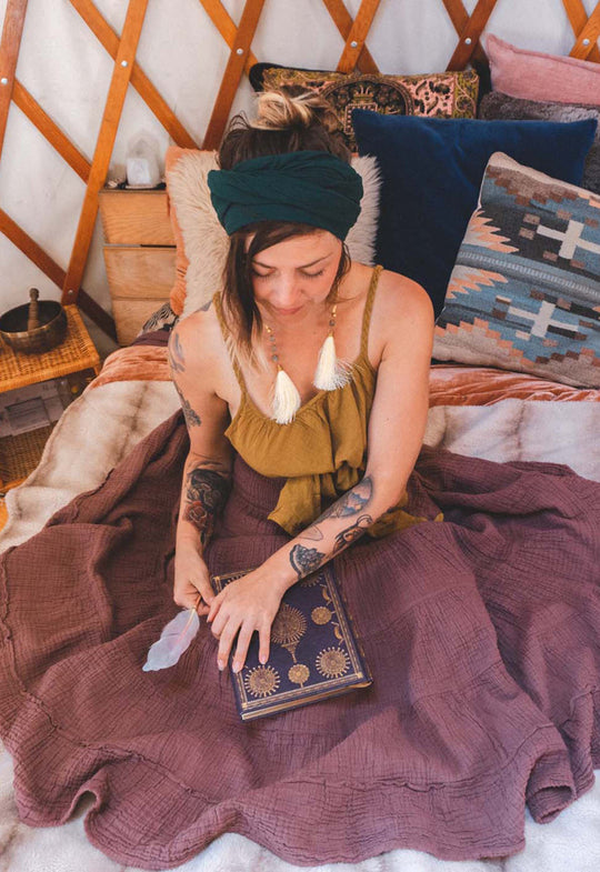 Woman sits on bed with hair wrapped in head scarf, tank top and long flowing skirt.