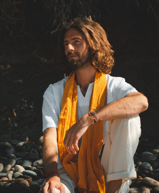 Man sits on rocks dressed in a white tshirt and natural pants with gold accent scarf. His left arm rests on bend left knee.
