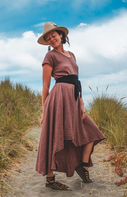 Female model is wearing brown hat and long purple gauze maxi dress tied at waist with black sash. She is posed outside in the dunes by the ocean wearing sandals.