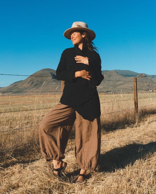 Woman is posed near wire fence in long brown balloon pants and black wrap.