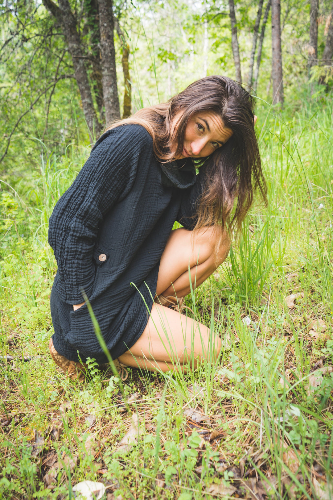 Model kneels in green grass wearing a long black gauze shirt with long sleeves. Her legs are bare.