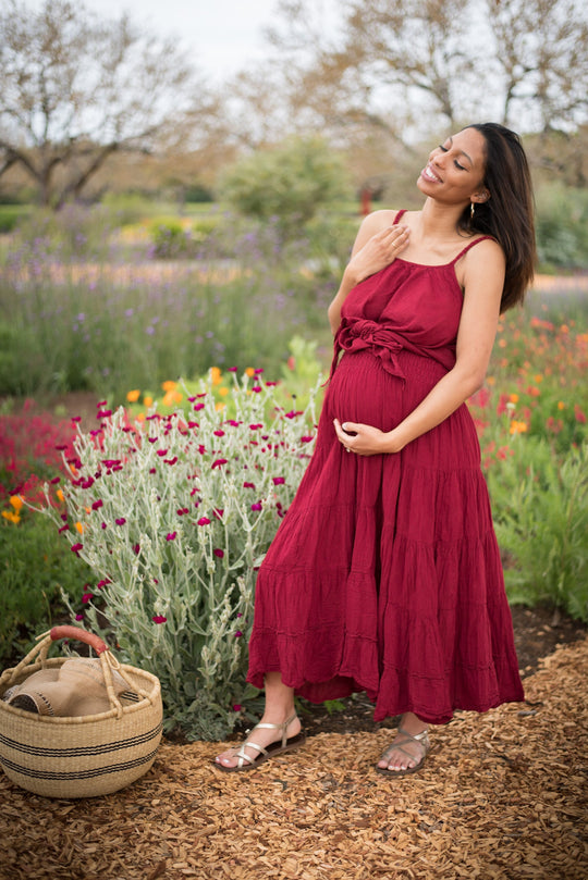 Female model wears maternity clothing and is holding her pregnant belly while in a field of flowers. She is wearing a ruby red tank top, ruby red maxi skirt and silver sandals. There is basket at her feet.