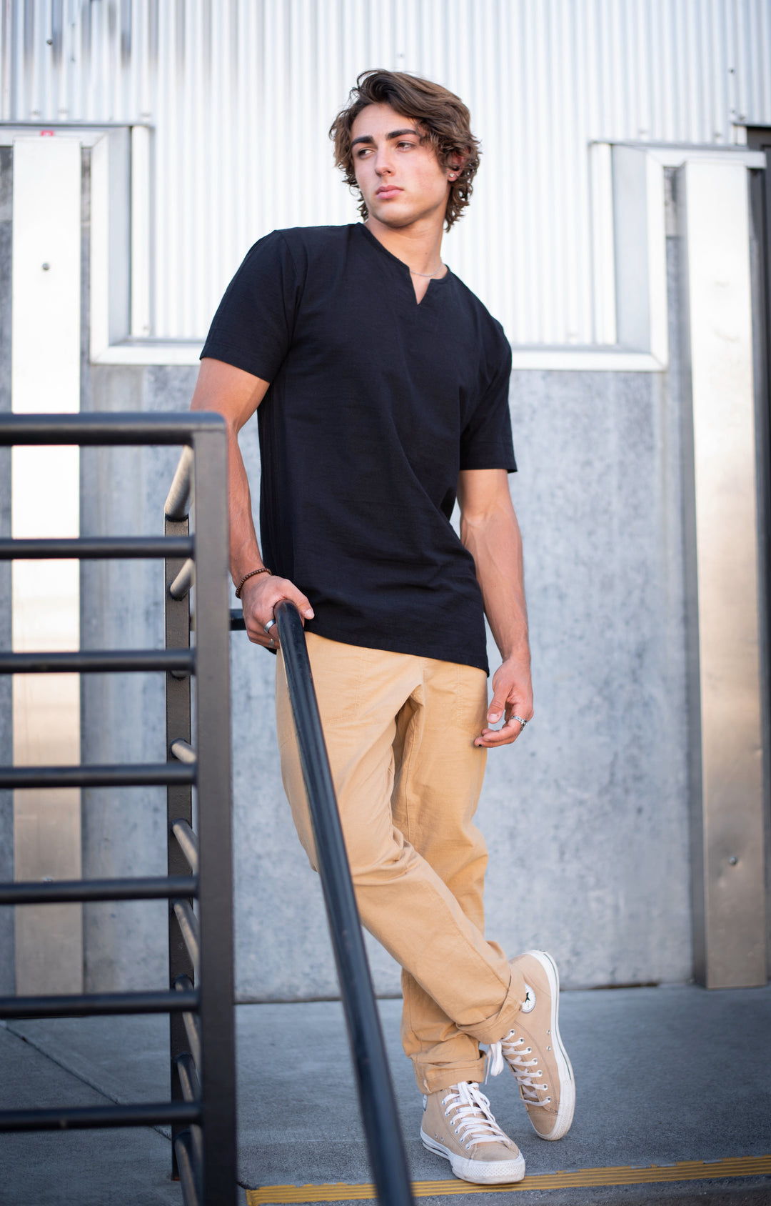 Male model is wearing black shirt and tan pants with laced sneakers. He leans agains a railing in a stairway.