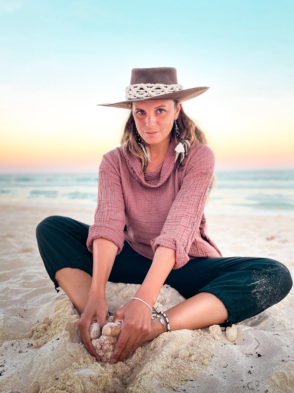 Female model is clothed in pink shirt with long sleeves and navy leggings. She sits on a sandy beach with ocean in behind her.