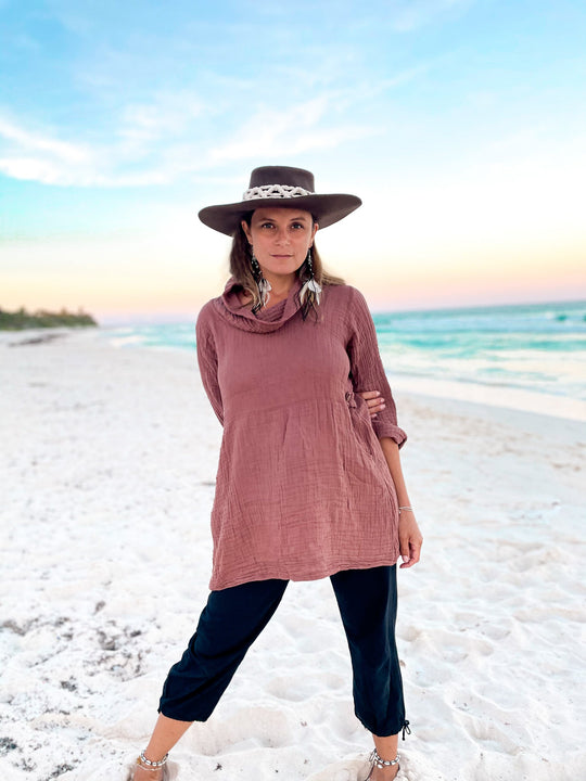 Female model stands on sandy beach in a hat, pink cowl sweater and navy drawstring pants.