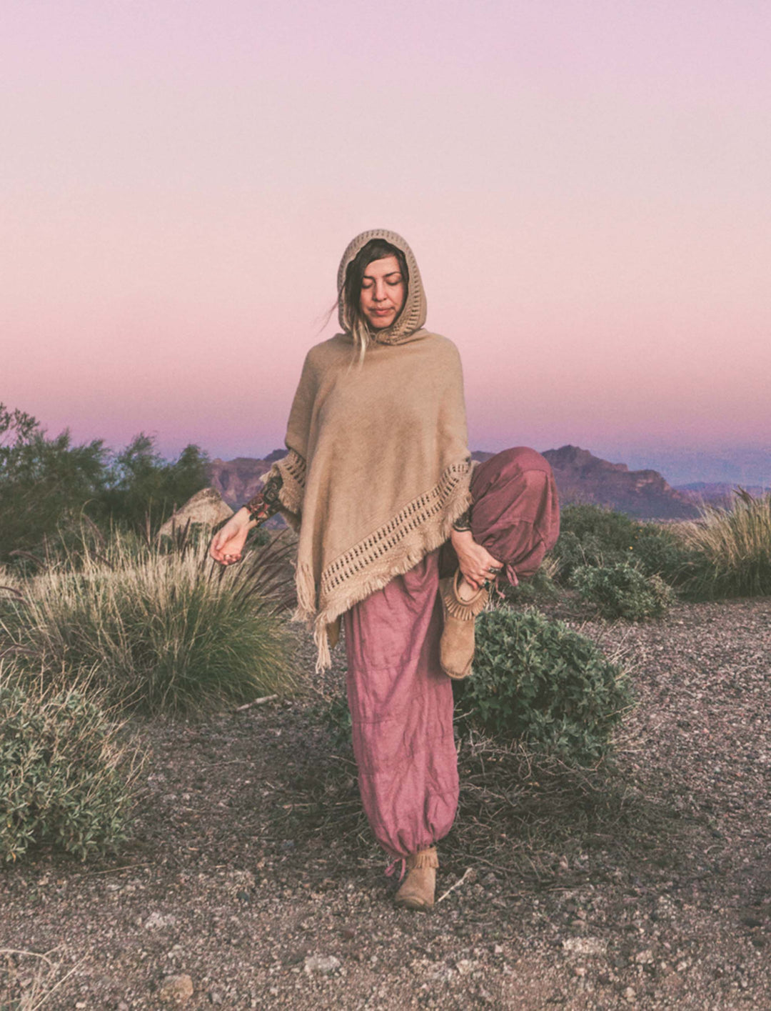 Female in tree pose wears pink gauze balloon pants with knit brown poncho.