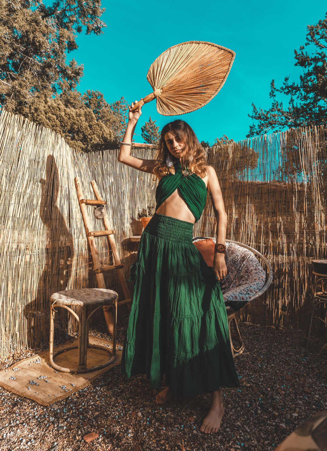 Female model is clothed in emerald halter top and tiered skirt. She holds a reed fan above her head.